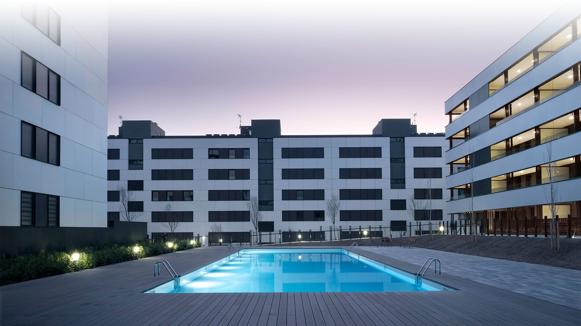 Edificios de viviendas alrededor de una piscina iluminada al anochecer.